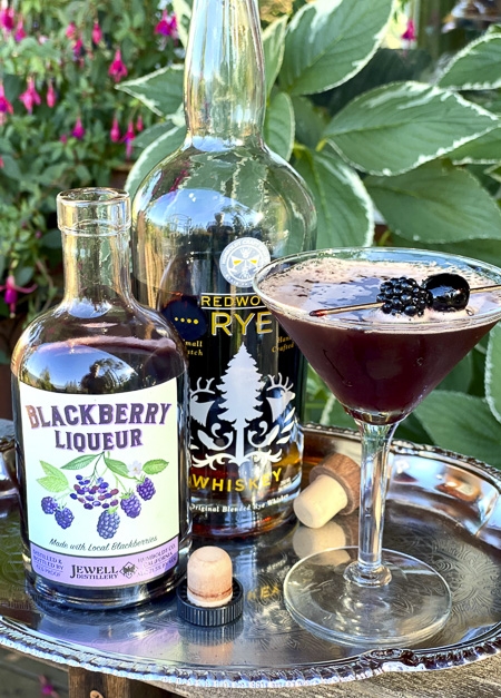 Blackberry liqueur and a bottle of rye whiskey with a martini glass garnished with a blackberry and luxardo cherry