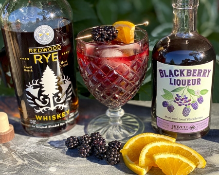 Blackberry liqueur bottle and a rye whiskey bottle with an old fashioned glass filled with a cocktail and garnished with blackberries and an orange slice